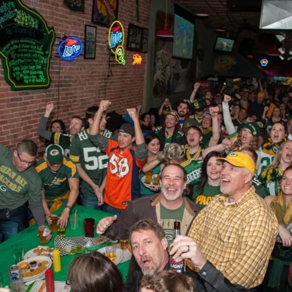 Excited crowd of Green Bay Packers fans cheering in a lively bar-and-grill, with food and drinks.