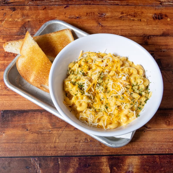 A white bowl of creamy macaroni and cheese with shredded cheese and parsley, served with toast.