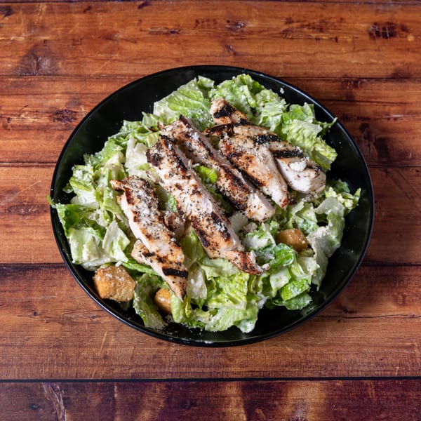 Grilled chicken Caesar salad with croutons and parmesan cheese in a black bowl on a wooden table.