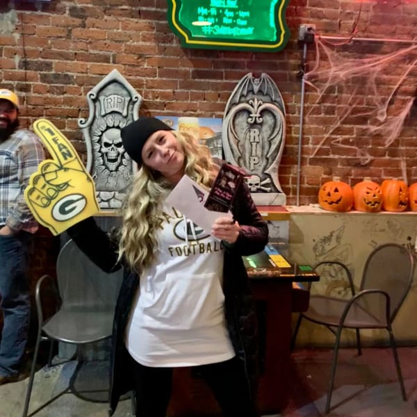 A woman in a Packers shirt and foam finger poses in a bar with Halloween decorations.