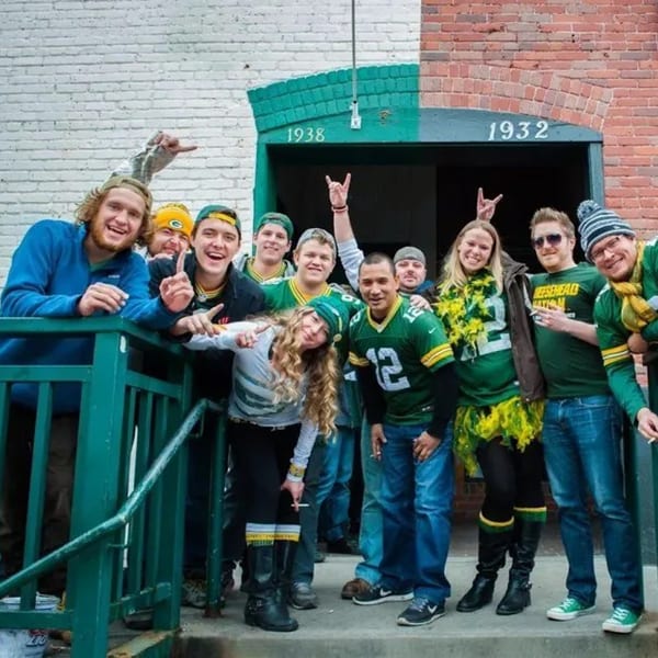 A group of friends in green and gold Packers gear pose happily outside Swanky's Vittles and Libations.