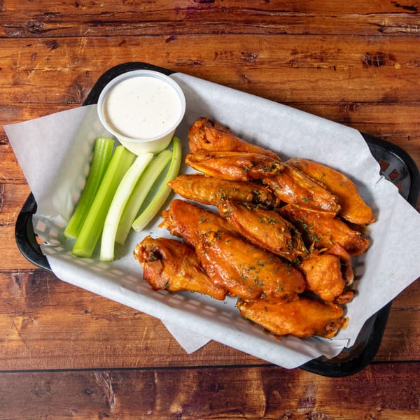 A basket of Swanky's buffalo wings with celery sticks and ranch dressing on a wooden table.