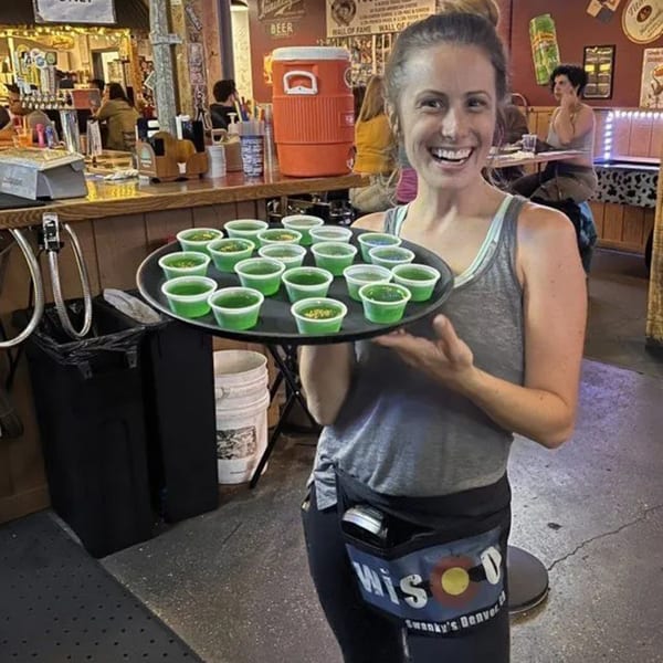 Smiling Swanky's server holding a tray of green jello shots in a lively bar setting.