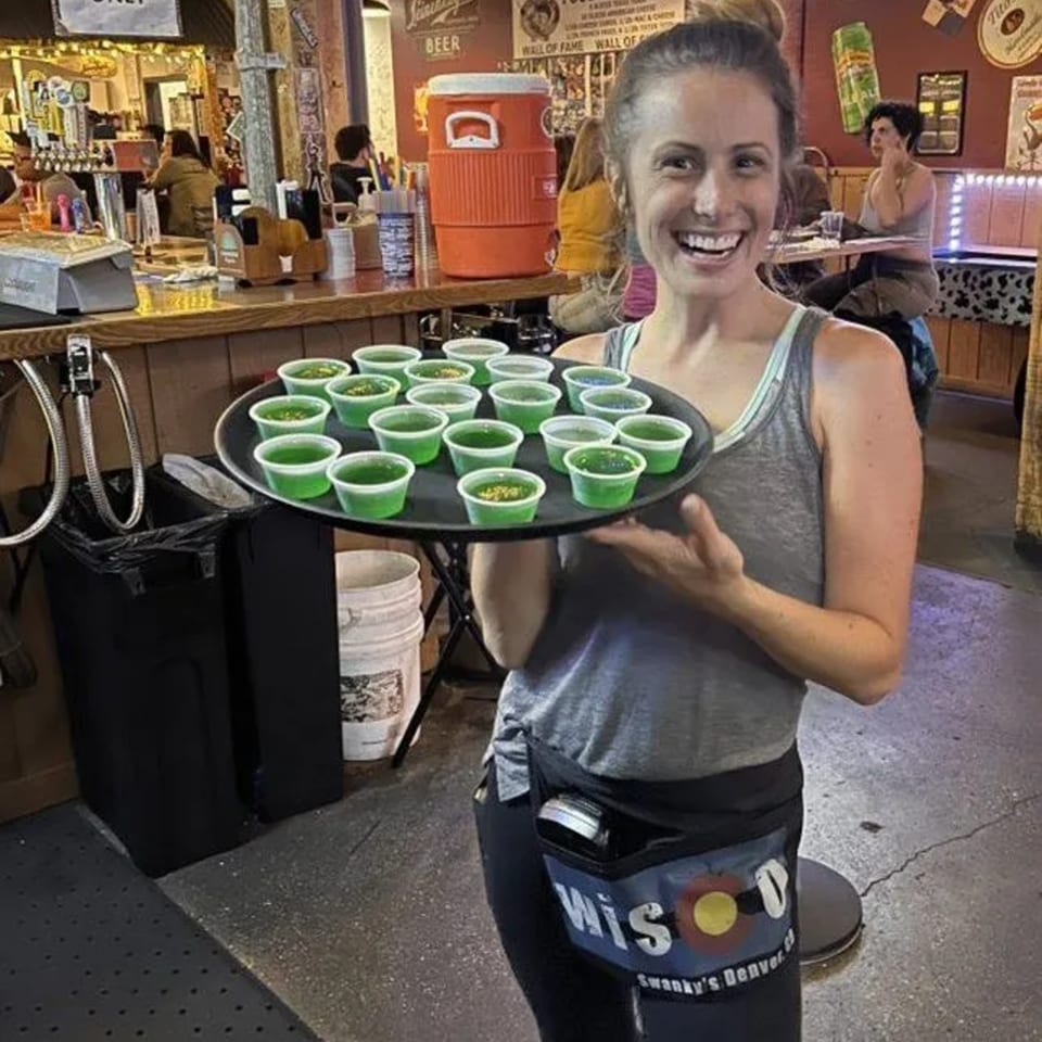 Smiling Swanky's server holding a tray of green jello shots in a lively bar setting.