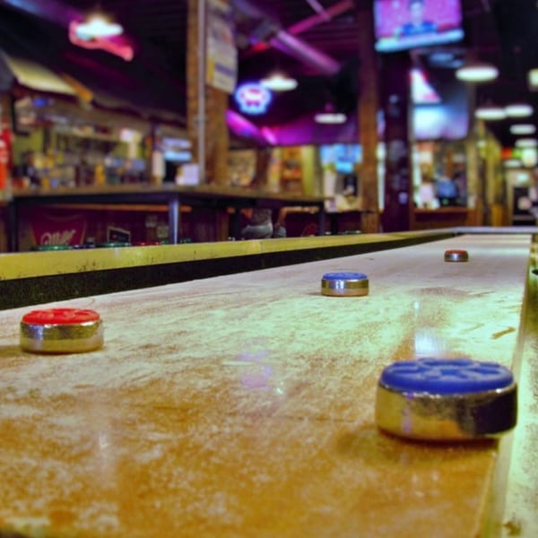 Shuffleboard table with red and blue pucks, in a dimly lit bar-and-grill setting.
