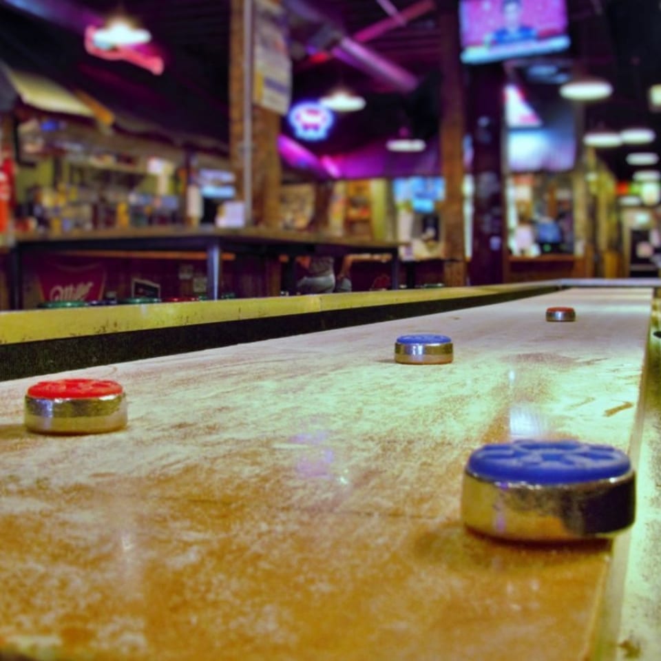 Shuffleboard table with red and blue pucks, in a dimly lit bar-and-grill setting.